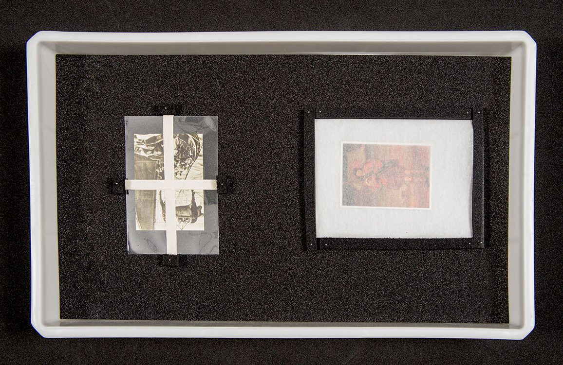 Two paper objects packed in plastic envelopes. They are both sitting in a plastic box supported by black foam.